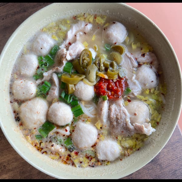 A close-up of a bowl of Thai noodle soup with meatballs, sliced pork, green onions, and chili paste.