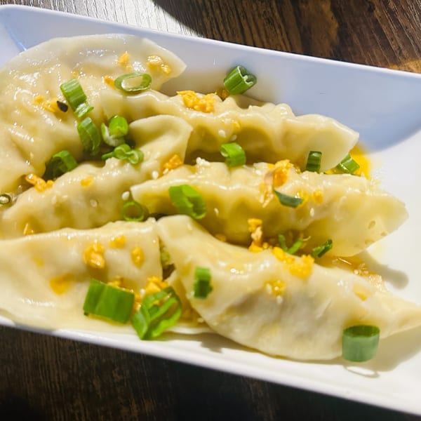 Steamed dumplings topped with chopped green onions, fried garlic, and sesame seeds on a white plate.