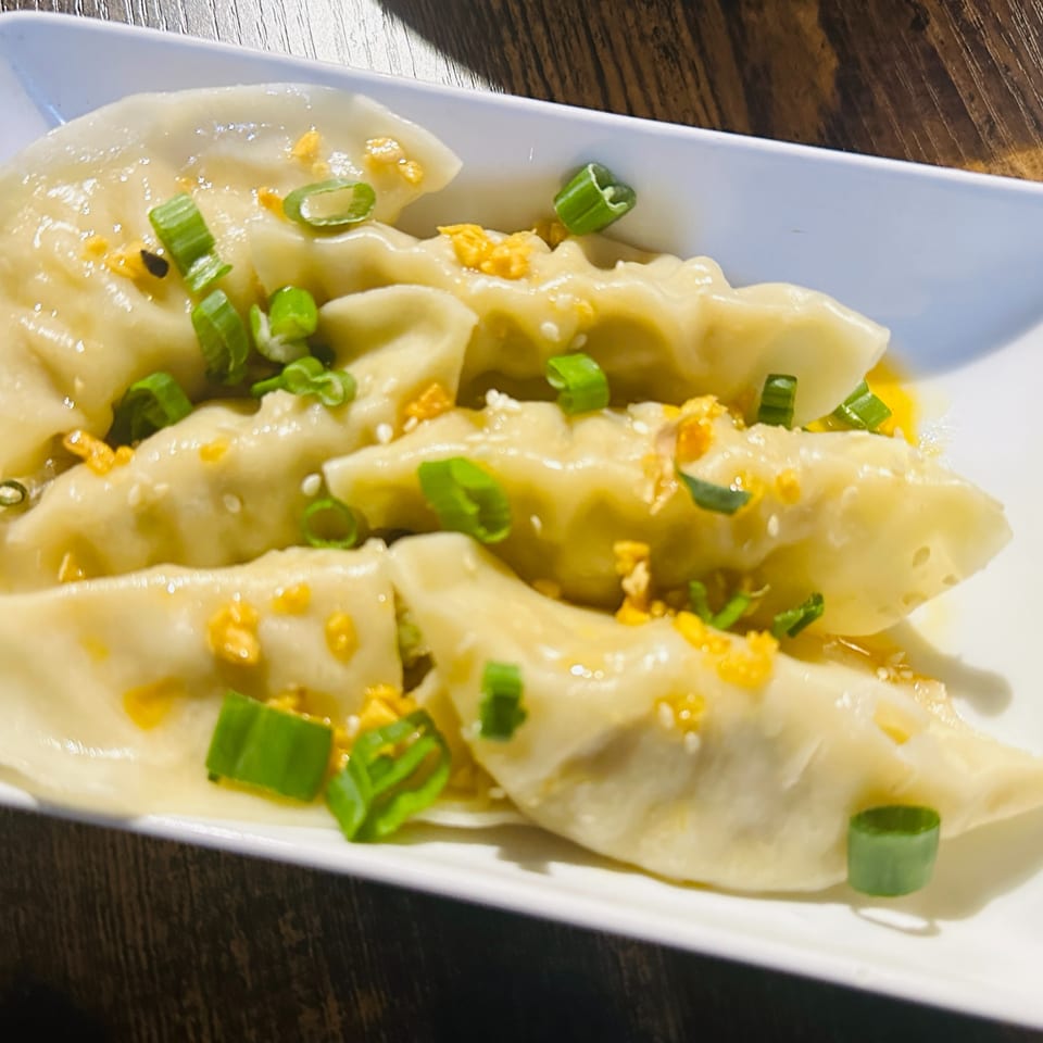 Steamed dumplings topped with chopped green onions, fried garlic, and sesame seeds on a white plate.