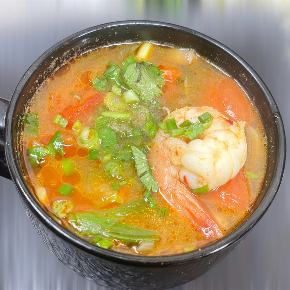 Close-up of a vibrant Tom Yum soup with shrimp, tomatoes, and fresh herbs in a black bowl.