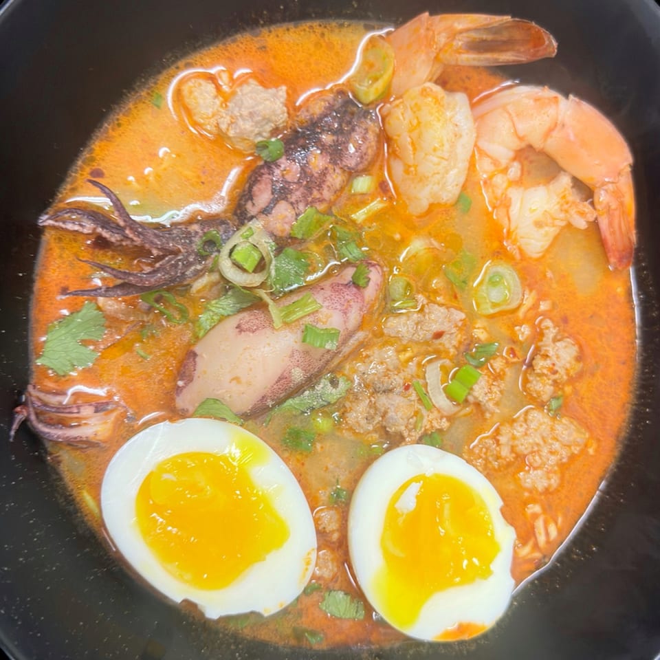 Spicy Thai ramen with shrimp, squid, ground pork, and two soft-boiled eggs in a black bowl.