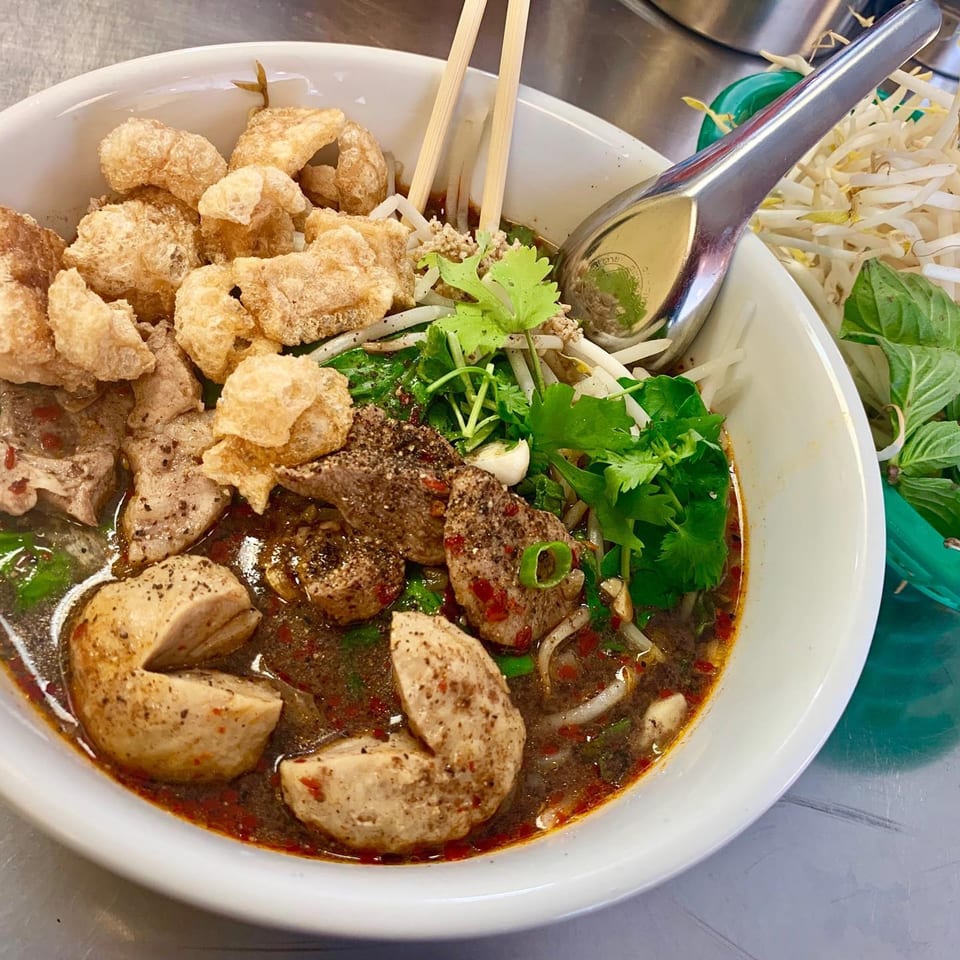 A close-up of a bowl of Thai boat noodles with pork, meatballs, and crispy pork rinds.