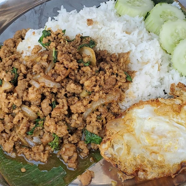 Thai basil chicken with rice, a fried egg, and sliced cucumbers on a metal plate.